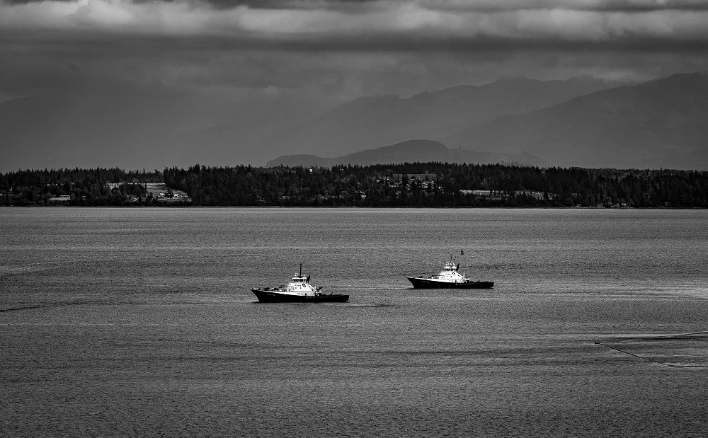 Two boats are sailing on calm waters with a forested shoreline and mountains visible in the distance under a cloudy sky.