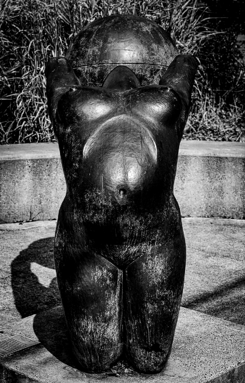 A bronze sculpture of a kneeling figure holding a large spherical object above its head is displayed outdoors with grass and a concrete bench in the background.