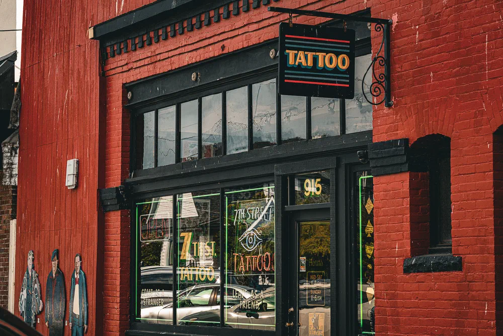 A tattoo shop with a black and orange sign is located in a red brick building, featuring large windows and artwork on the adjacent wall.
