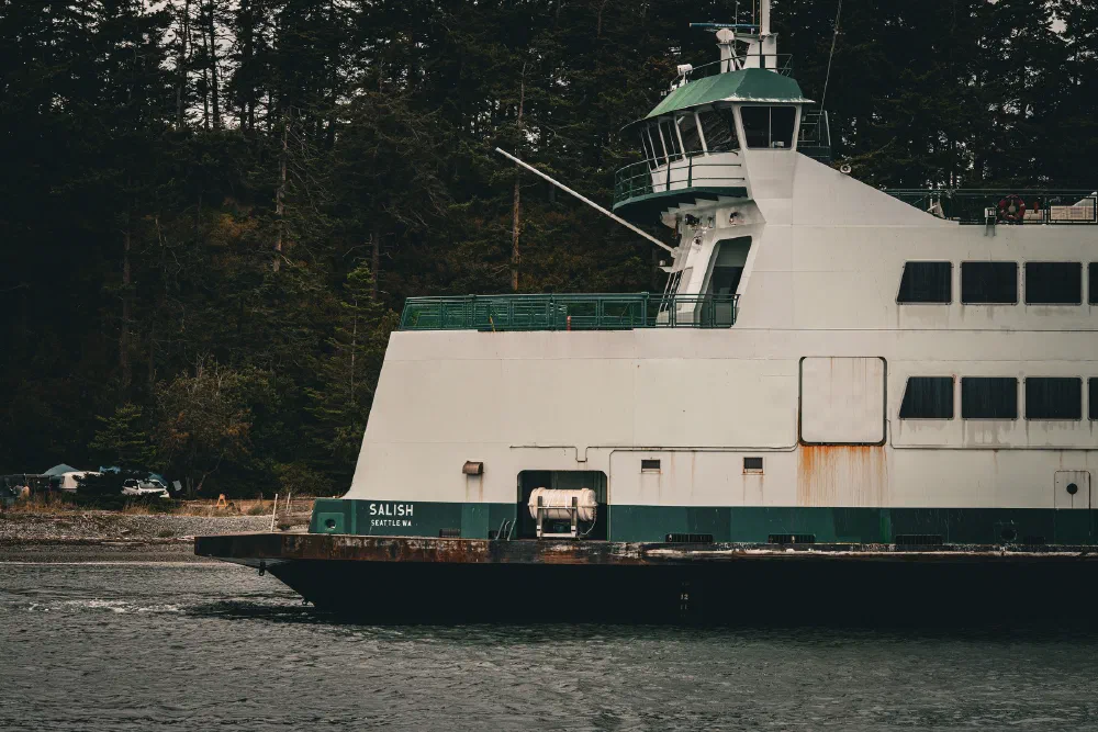 A large ferry named Salish is cruising on the water near a forested shoreline.