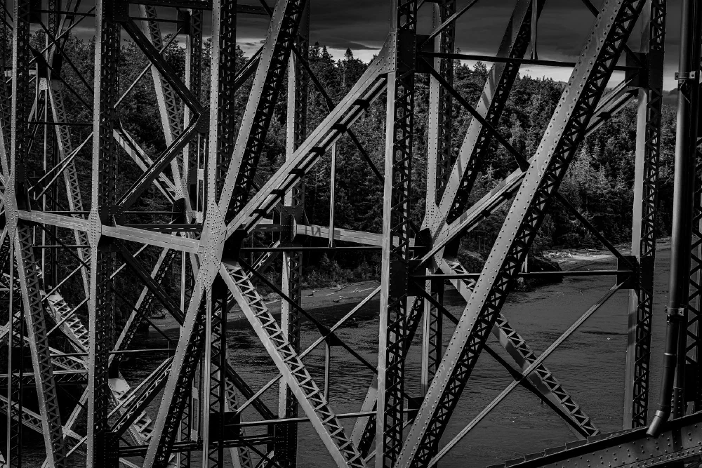 A detailed close-up of the intricate metal framework of a bridge shows a river and forested area in the background.