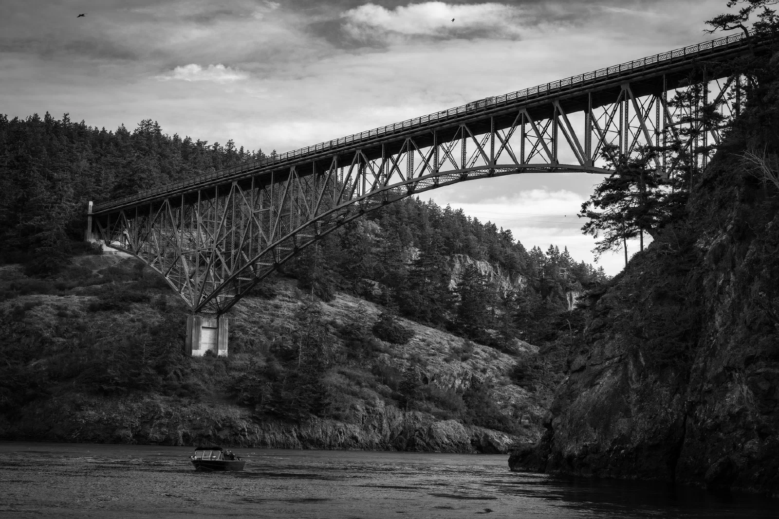 A large steel truss bridge spans across a river with forested hills on either side under a partly cloudy sky.