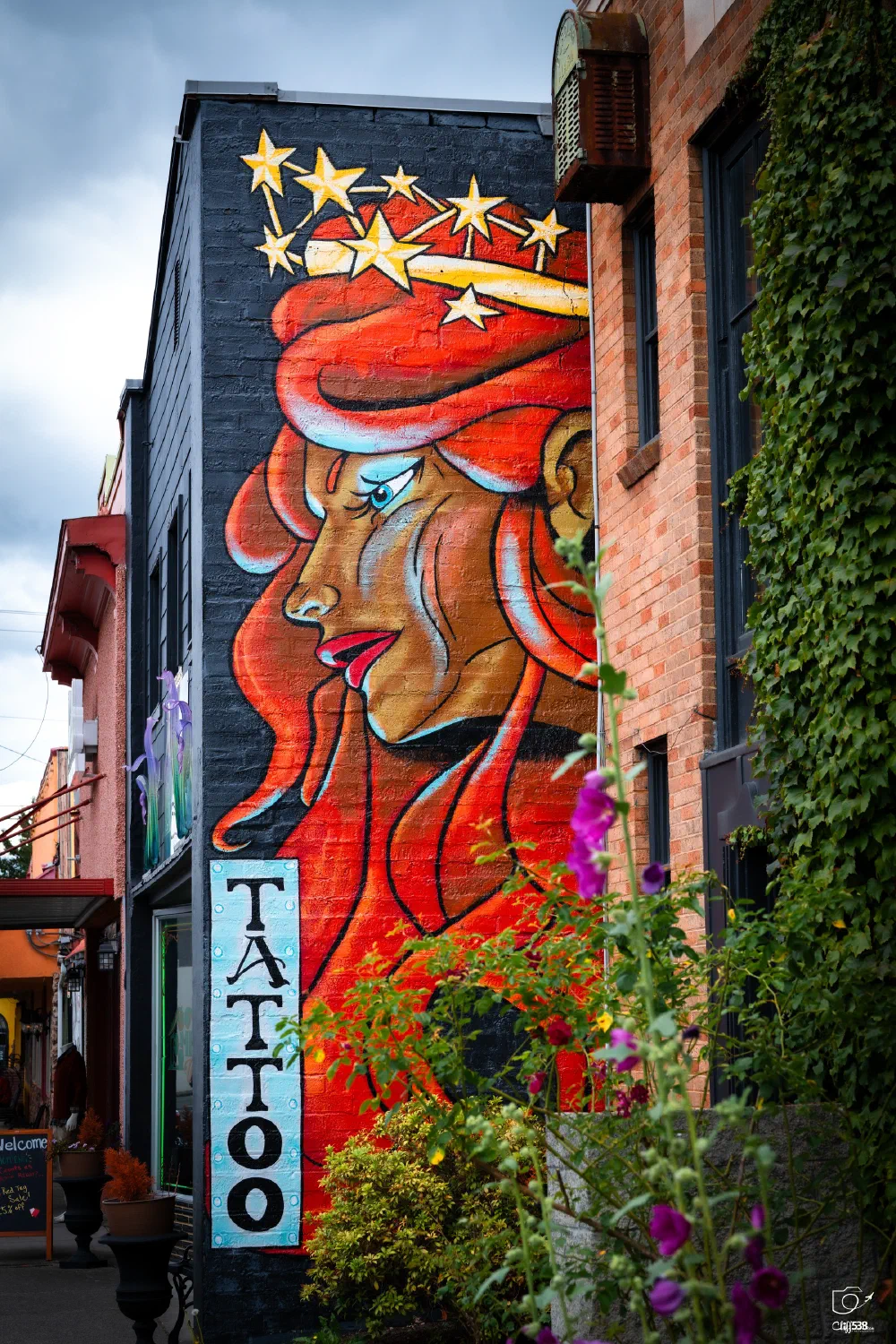 A vibrant mural of a woman with flowing red hair and a crown of stars adorns the side of a tattoo shop building.