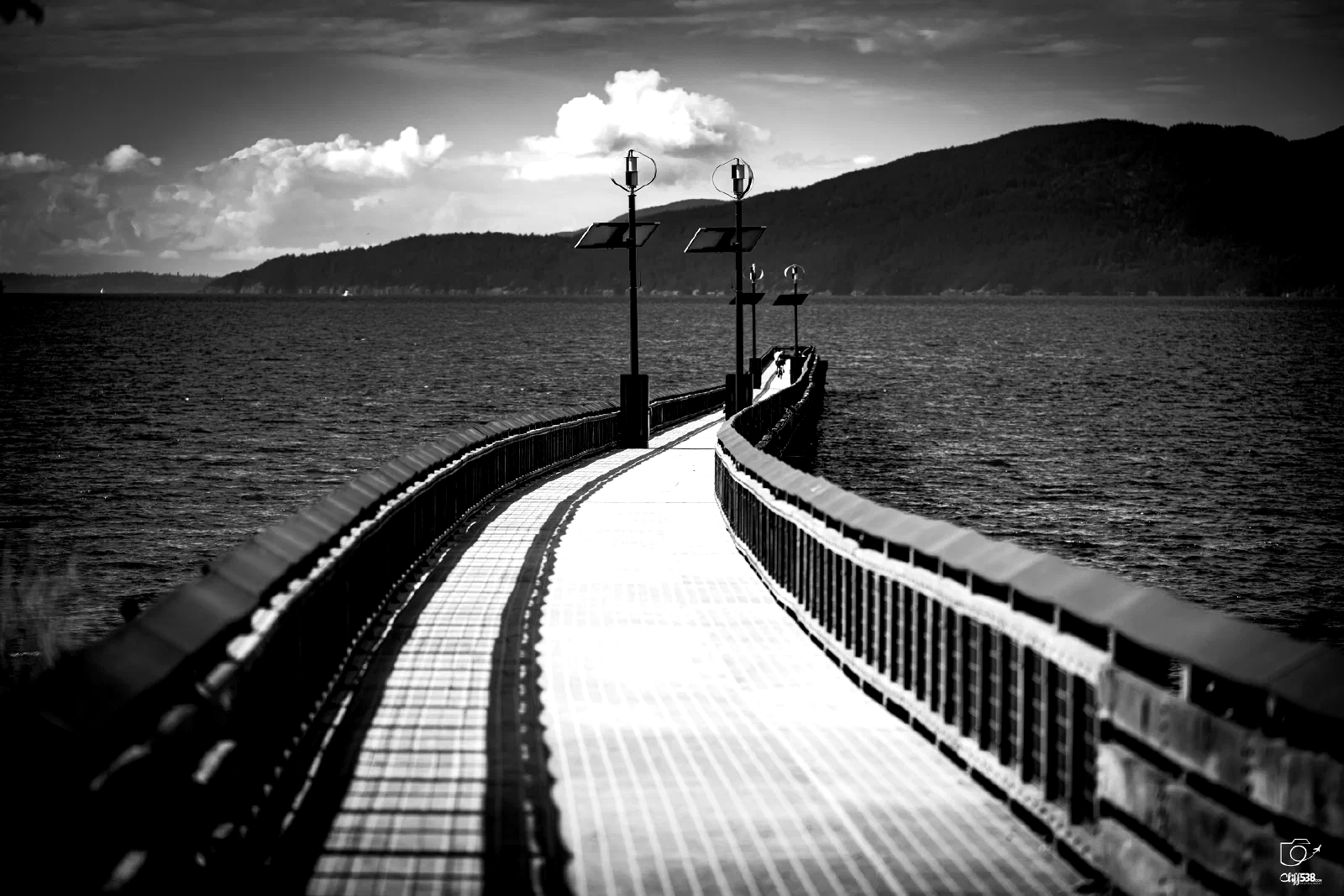 A curved wooden pier extends over a body of water towards a distant mountainous shoreline under a partly cloudy sky.