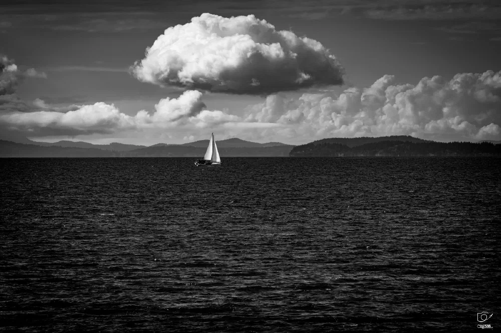 A lone sailboat glides across calm waters with a dramatic cloudy sky and distant landforms in the background.