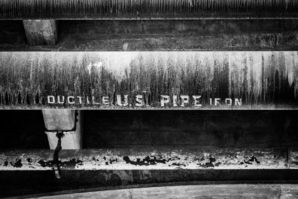 A weathered and rusted pipe labeled DUCTILE U.S. PIPE IRON is shown in a black and white photograph.