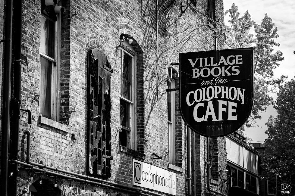 A brick building features signs for Village Books and the Colophon Cafe against a backdrop of trees and sky.