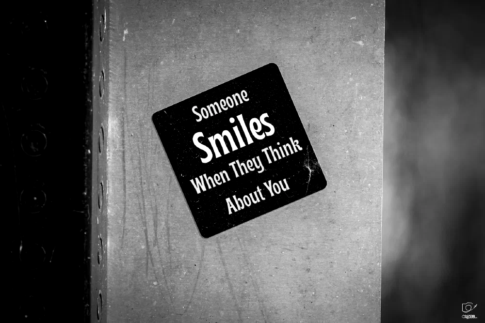 A black sticker with white text saying Someone Smiles When They Think About You is attached to a gray, textured surface.
