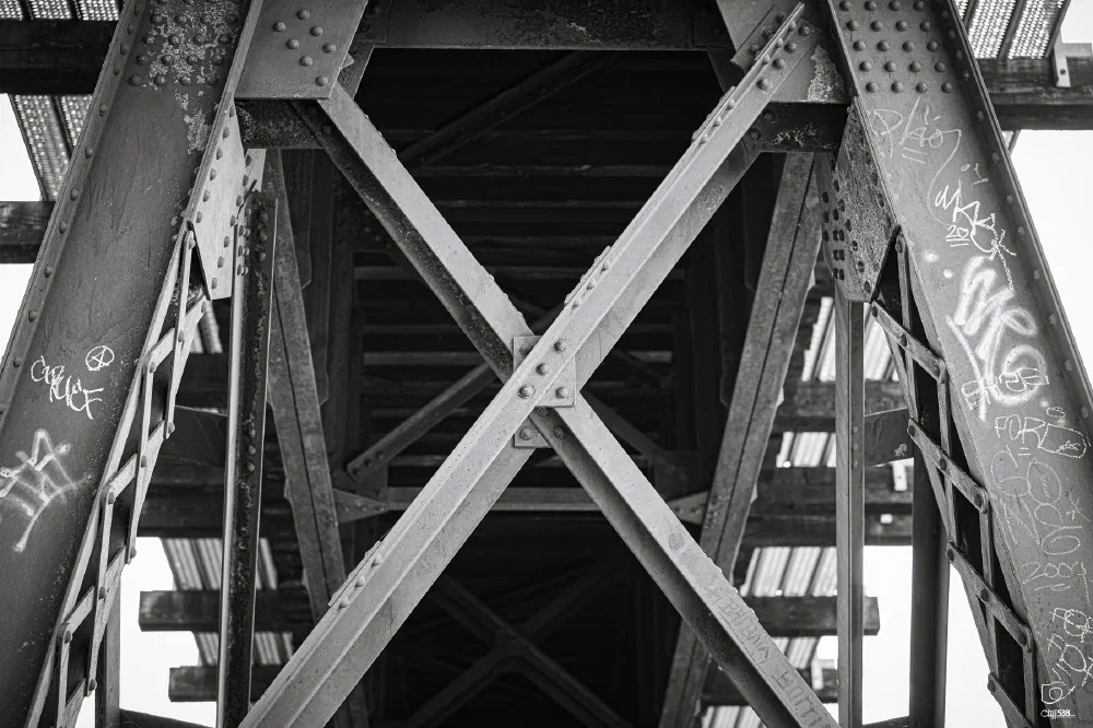 A black and white photo captures the underside of a metal bridge with graffiti on its support beams and riveted cross braces.