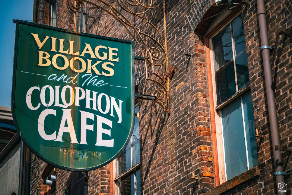 A green sign with yellow and white text hangs outside a brick building, advertising Village Books and The Colophon Cafe.