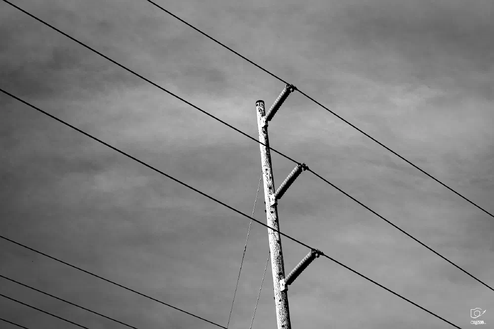 A wooden utility pole with multiple power lines stretches across a cloudy sky in a black and white photograph.