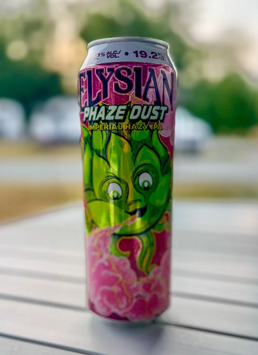 A colorful can of Elysian Phaze Dust Imperial Hazy IPA, featuring a green hop character and displaying an alcohol content of 9.2% and a volume of 19.2 fl oz, is placed on a white surface with a blurred outdoor background.