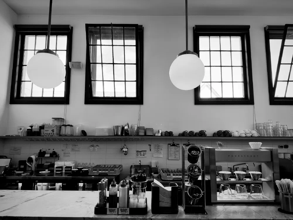A minimalist coffee shop counter features various coffee-making equipment, condiments, and three large pendant lights hanging below tall, gridded windows.