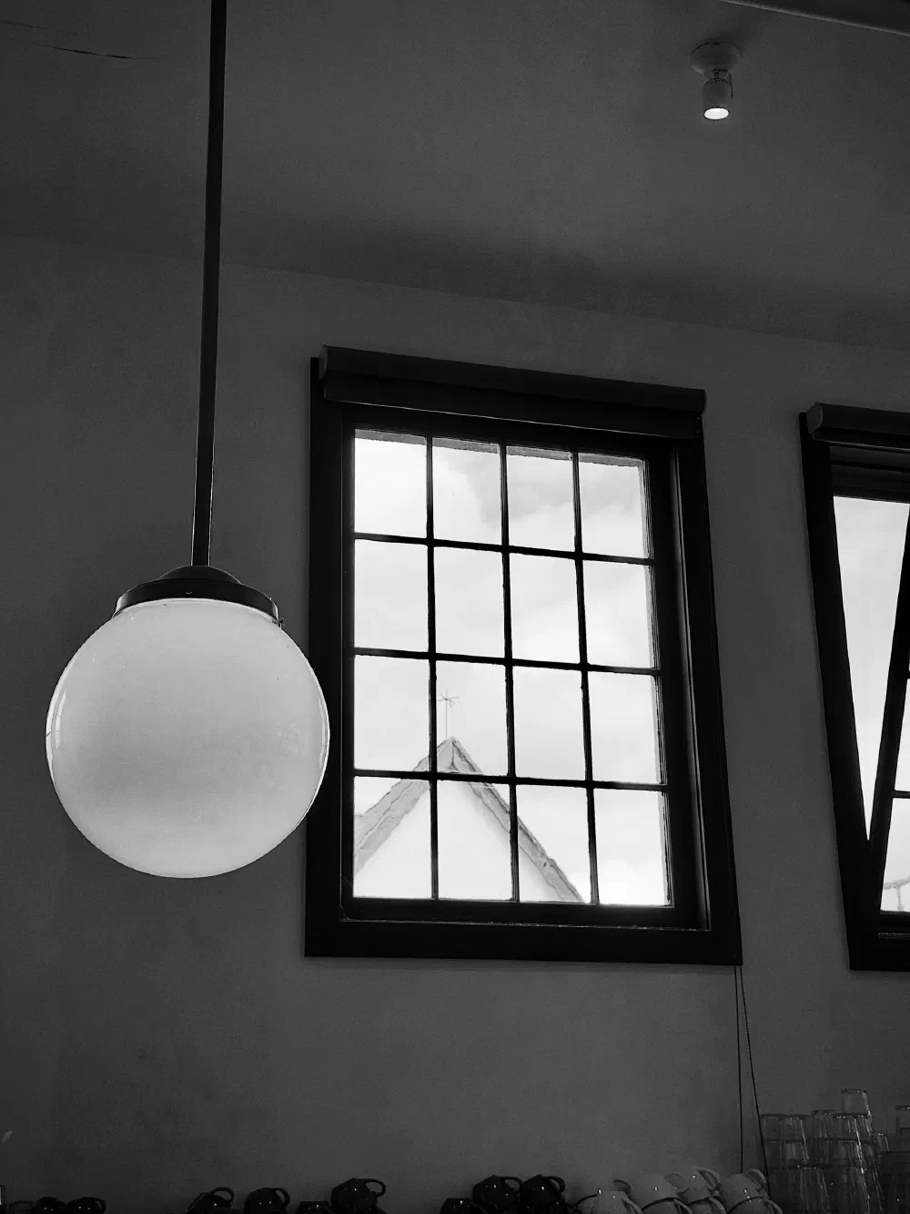 A round pendant light hangs near a window with a grid pattern, through which the peak of a roof and cloudy sky are visible.