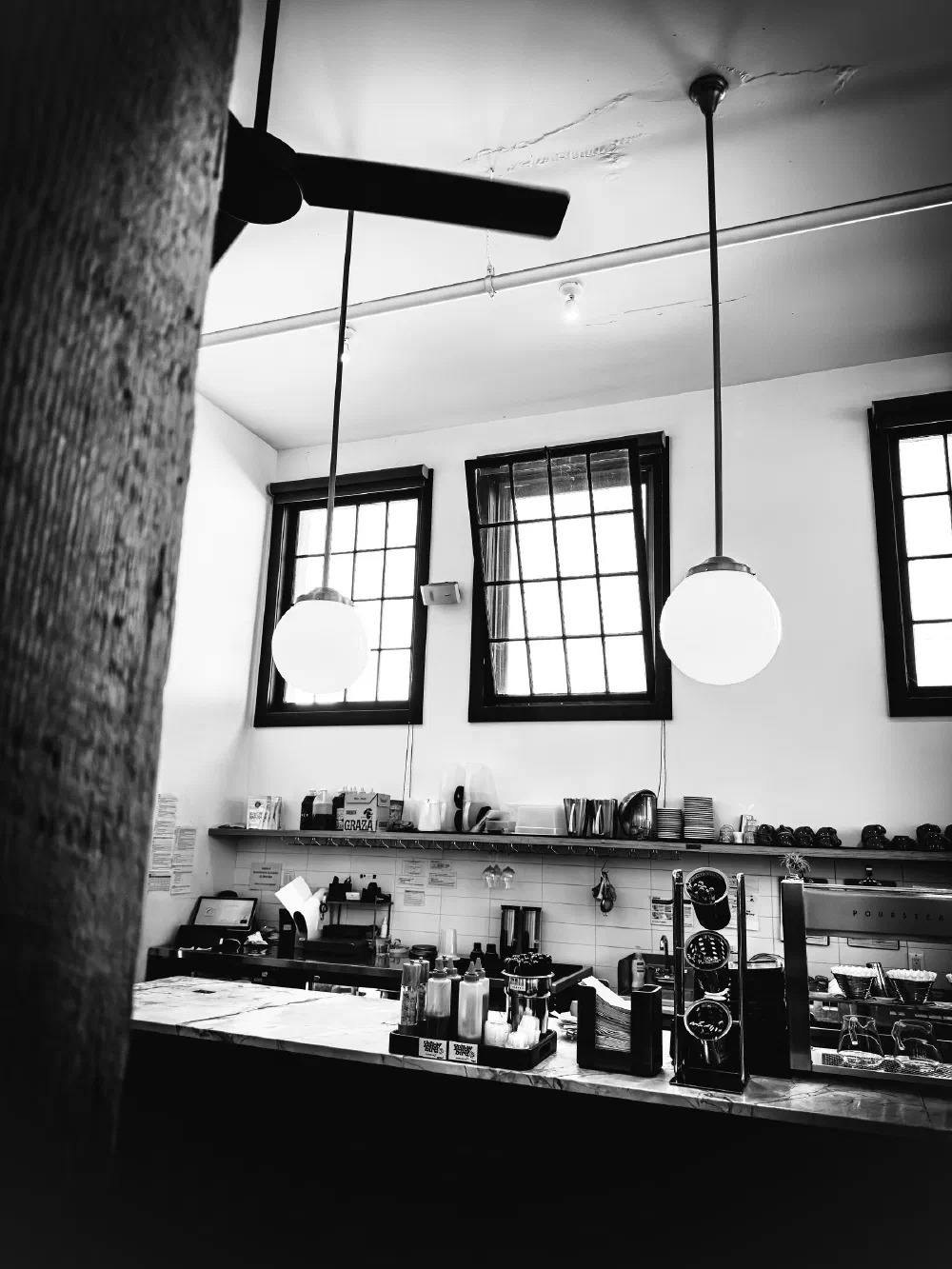 A black and white photo shows a coffee shop counter with hanging globe lights, windows, and various coffee-making equipment.