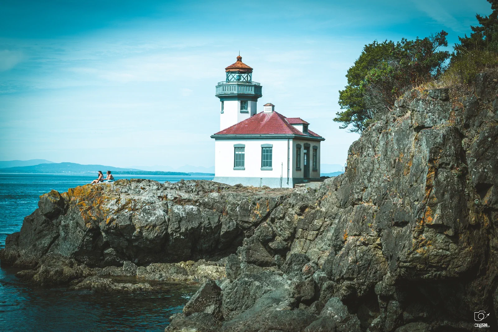 A lighthouse with a red roof stands on rugged cliffs by the sea under a clear blue sky.
