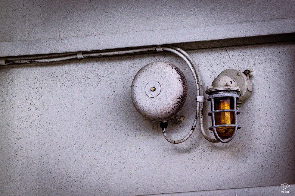 A rusty old fire alarm bell and a protective cage-covered light fixture are mounted on a worn white wall.