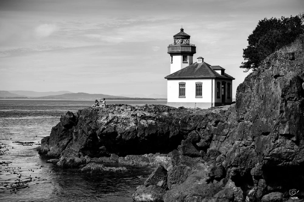 A lighthouse stands on a rocky cliff overlooking a calm body of water under a partly cloudy sky.