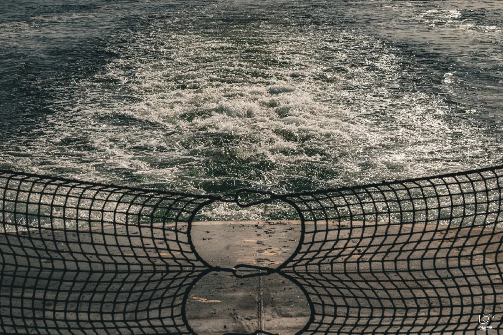 A view of turbulent water behind a boat, partially obscured by a safety net attached to the rear railing.