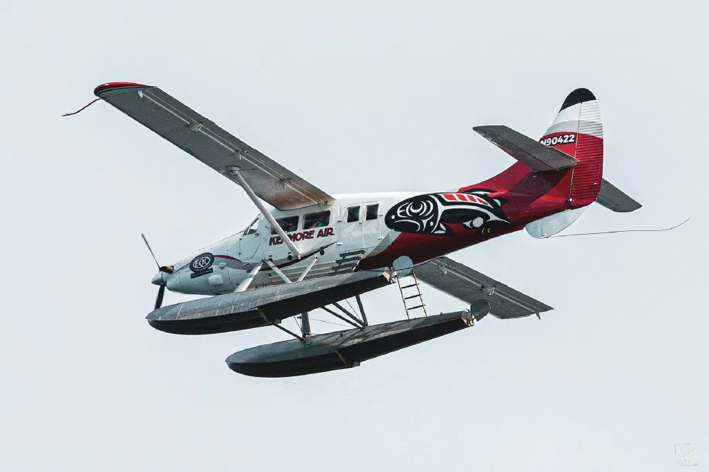 A floatplane with red, black, and white markings is flying against a clear sky.
