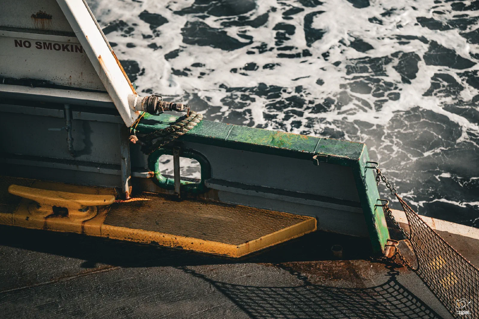 Part of a boat deck is seen with a yellow platform, green railing, and churning ocean water in the background.