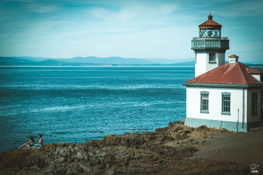 A lighthouse stands on a rocky coastline while two people sit near the water's edge overlooking the calm blue sea.