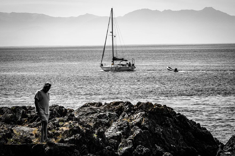 A person stands on rocky terrain near the shore while a sailboat floats on the calm sea with mountains visible in the distance.
