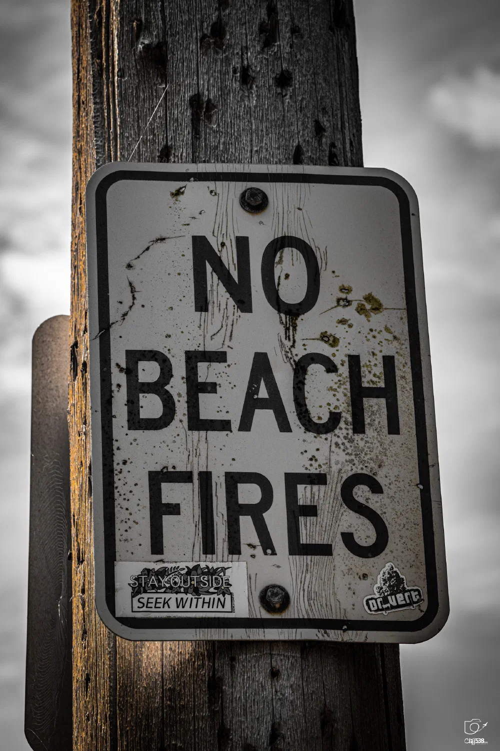 A weathered sign attached to a wooden post forbids beach fires and is marked with stickers, set against a cloudy sky.
