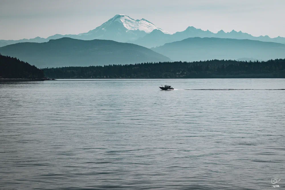 A boat is cruising across a calm body of water with a snow-capped mountain and forested hills in the background.
