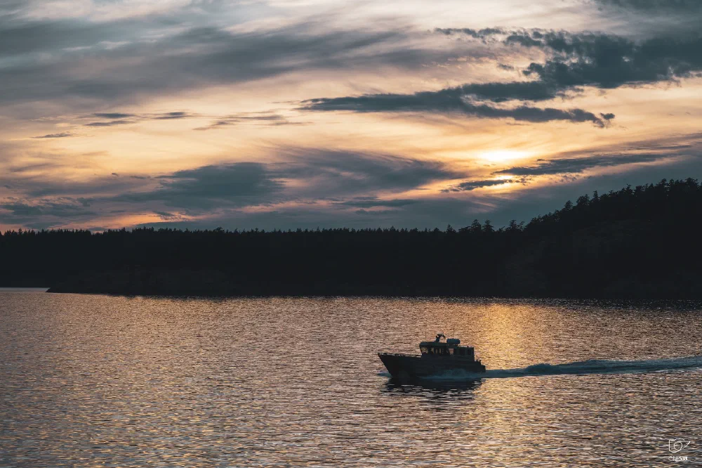 A boat is sailing across calm waters under a dramatic sunset sky with silhouetted trees on the horizon.