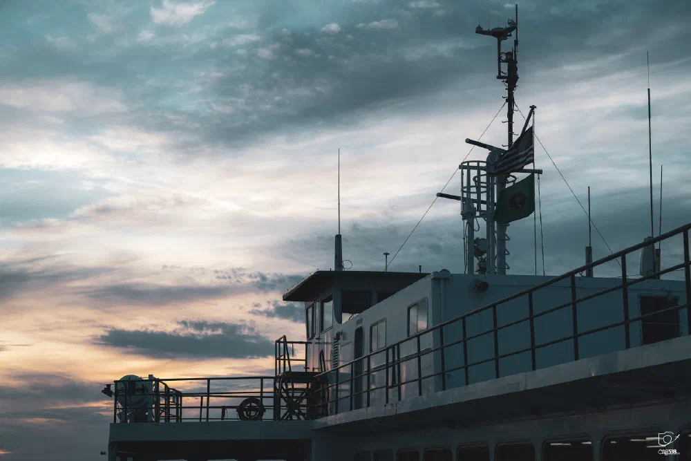 A boat or ship cabin with antennas and flags is set against a partly cloudy evening sky.