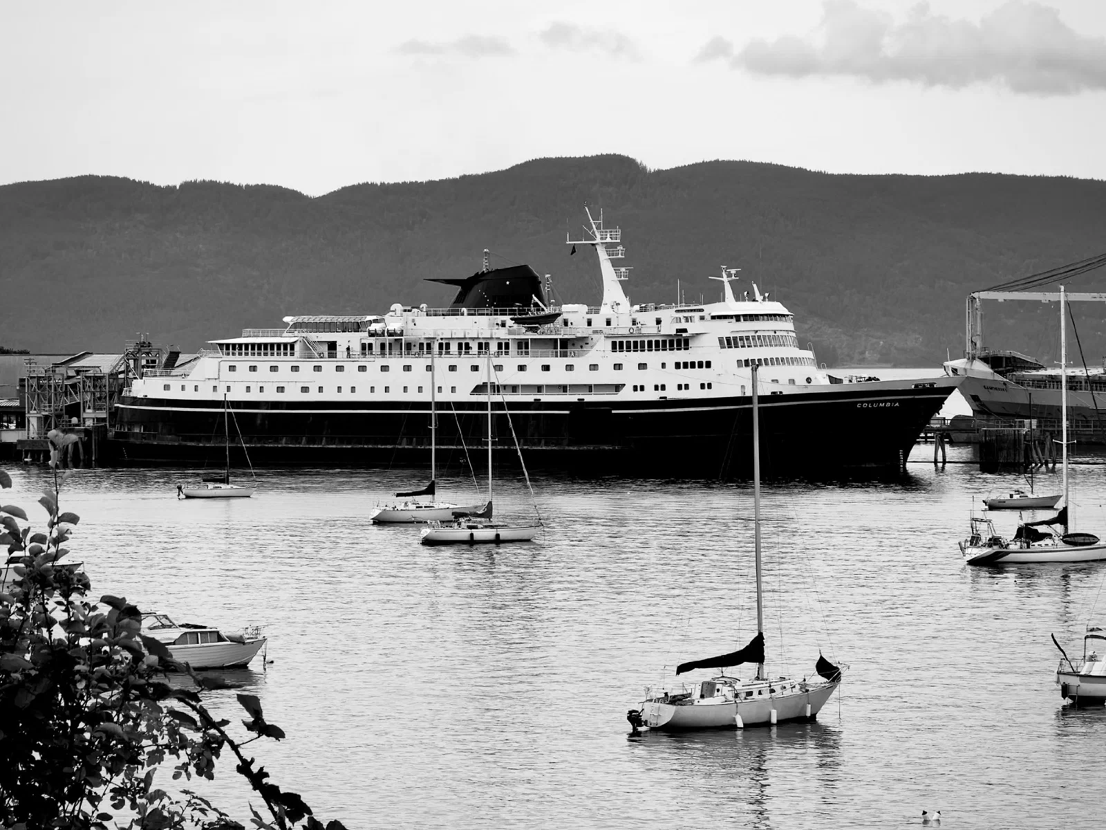 A large passenger ship is docked at a harbor with several smaller sailboats and watercraft floating nearby, set against a backdrop of hills.