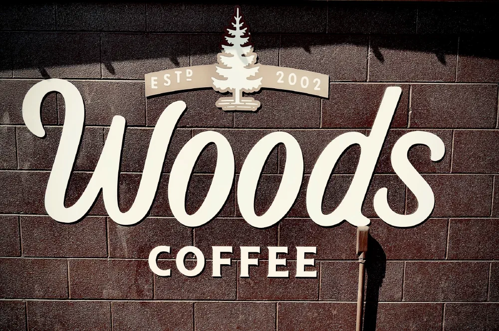 A sign for Woods Coffee, established in 2002, is displayed on a brick wall along with a tree graphic.