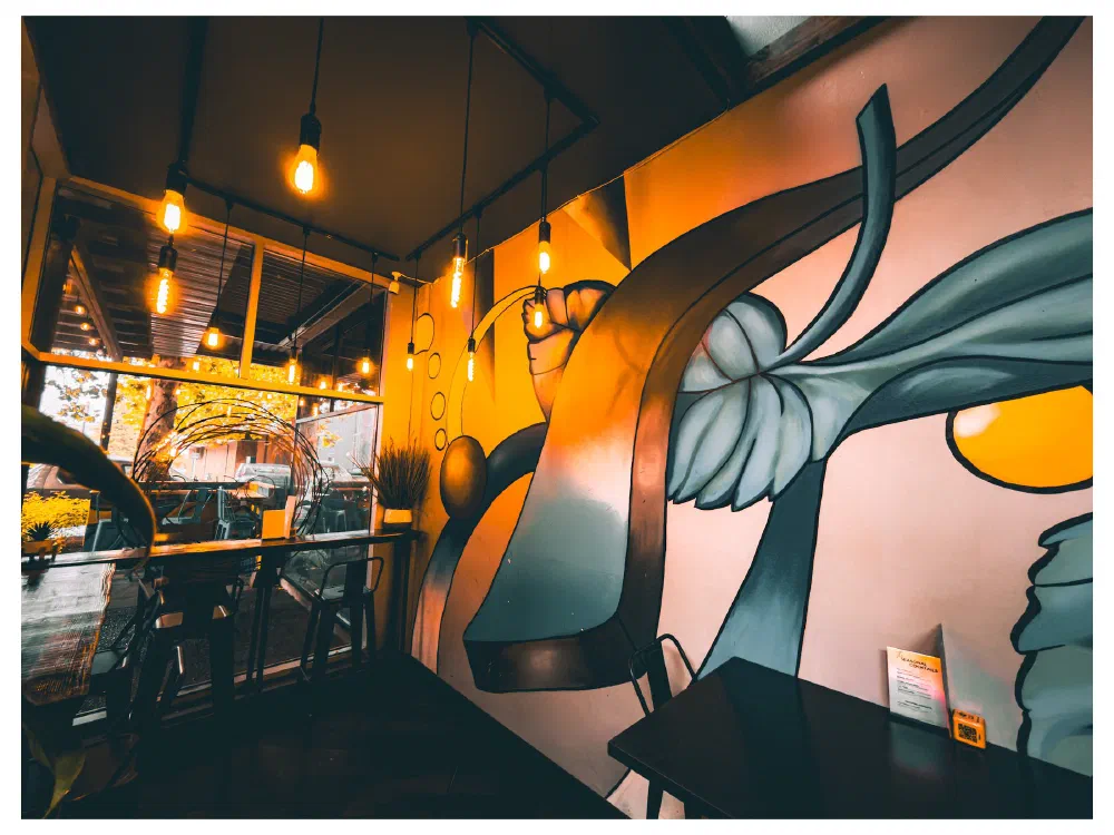 A cozy café corner features industrial-style hanging lights, wooden stools, a large window with a view outside, and a vibrant mural painted on the wall.