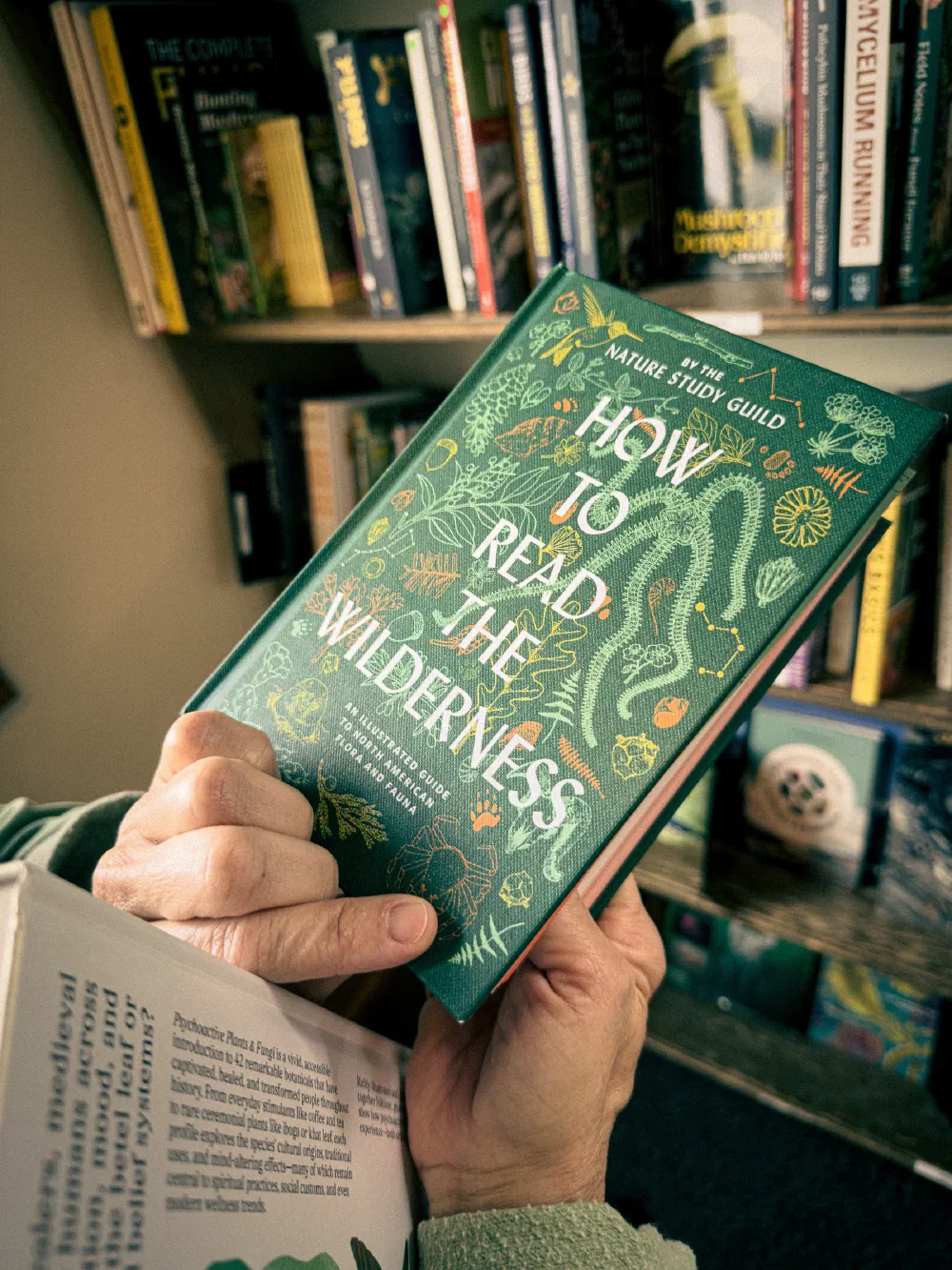 A person is holding an open book titled How to Read the Wilderness in front of a bookshelf filled with various nature and wilderness-related books.