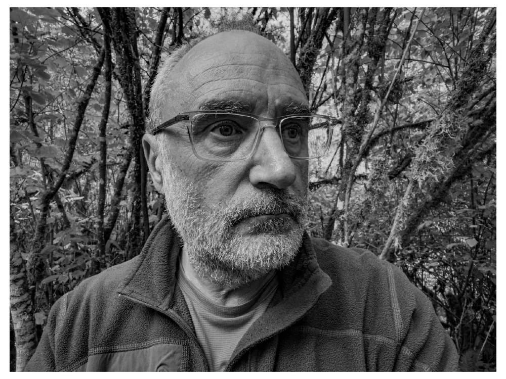 An older man with glasses and a beard is looking thoughtfully to the side while standing in a wooded area.