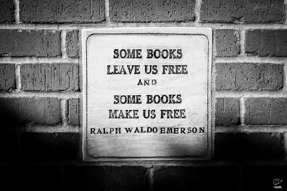 A plaque mounted on a brick wall displays a quote by Ralph Waldo Emerson: Some books leave us free and some books make us free.