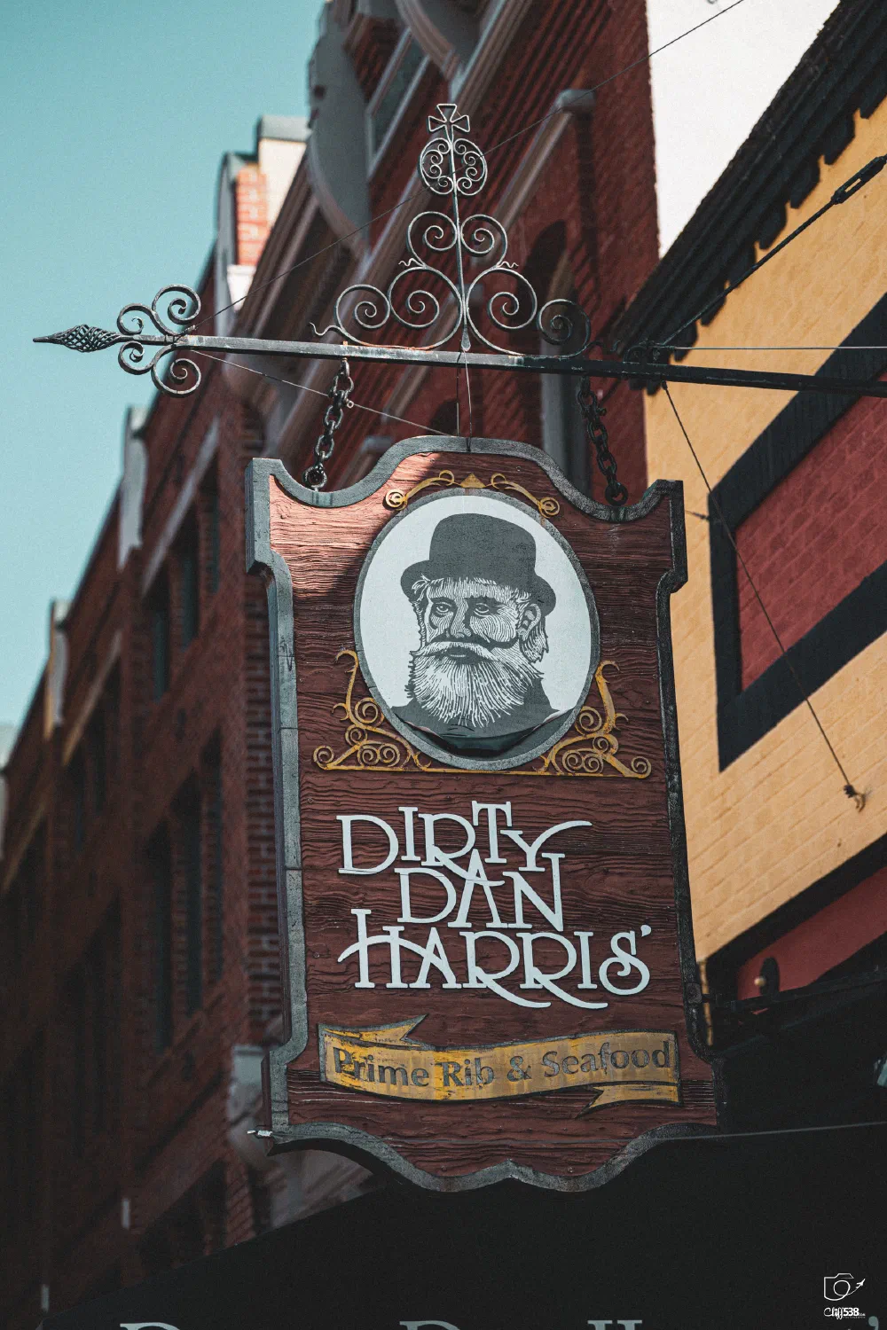 A wooden hanging sign for Dirty Dan Harris' featuring an illustrated bearded man in a top hat advertises prime rib and seafood outside a brick building.