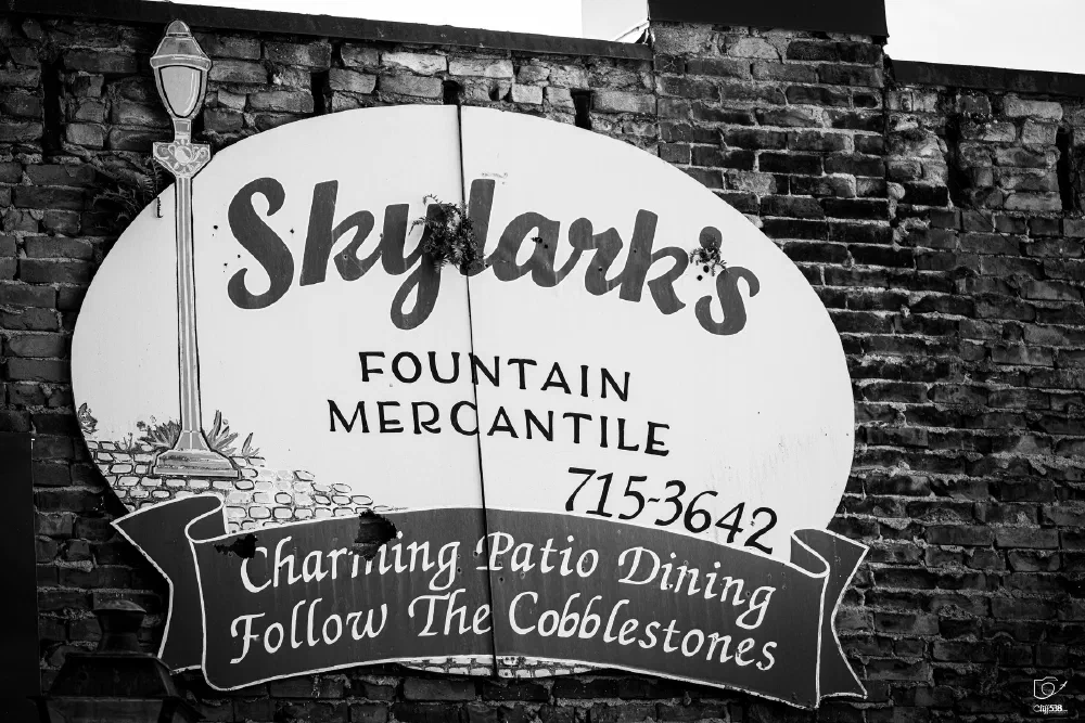 A vintage-style sign on a brick wall advertises Skylark's Fountain Mercantile and charming patio dining with directions to follow the cobblestones.