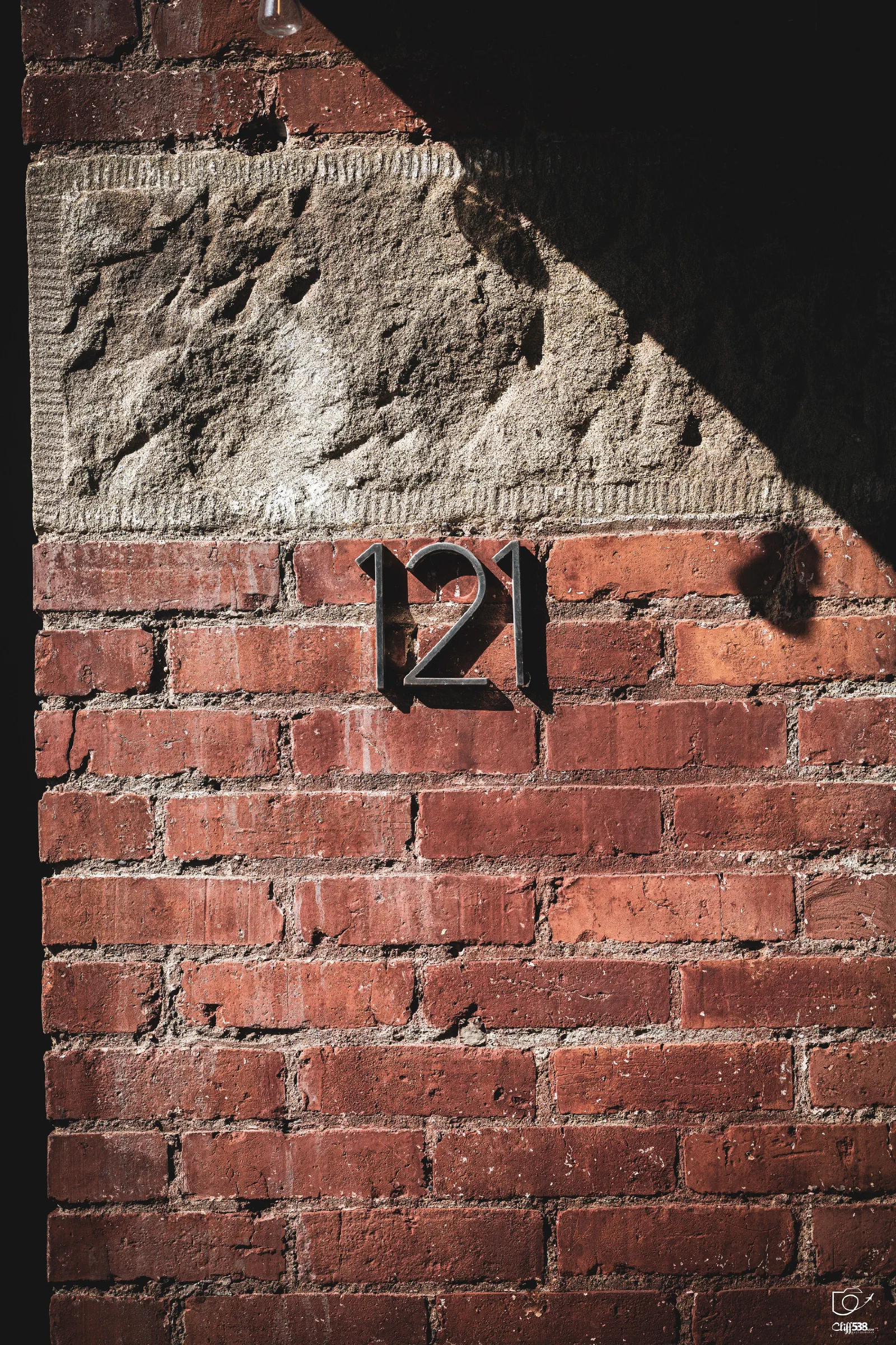 The number 121 is displayed on a red brick wall with a rough stone section above it, partially shaded by an overhead structure.