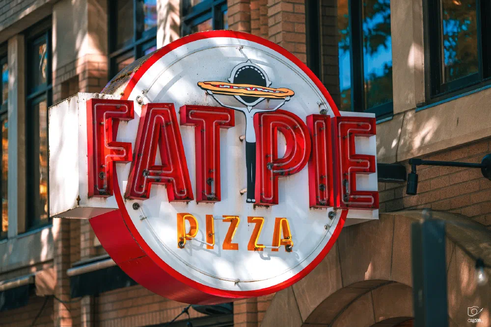 A brightly lit neon sign for Fat Pie Pizza is mounted on the side of a brick building.