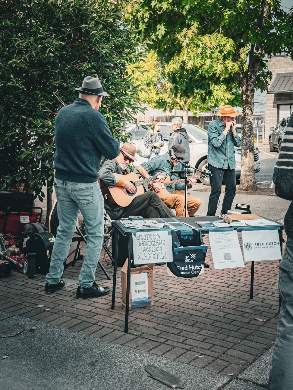 A group of musicians is performing outdoors near a table with signs supporting the Fred Hutch Cancer Center and a cause related to cancer awareness.