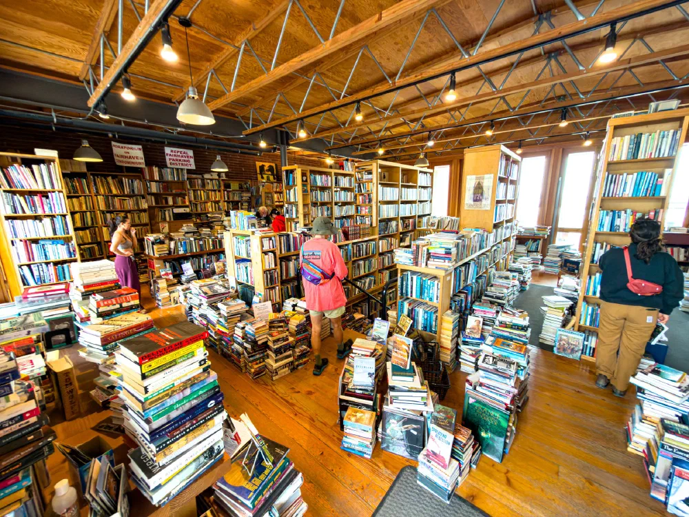 A bustling bookstore with tall wooden shelves and stacks of books on the floor, filled with several people browsing.