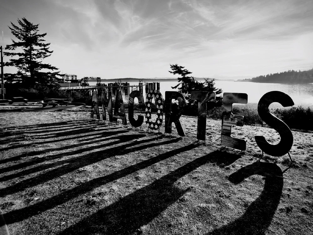 Large silhouetted letters spelling WALCARTES stand on a beach near a dock, casting long shadows in the light of the setting or rising sun.