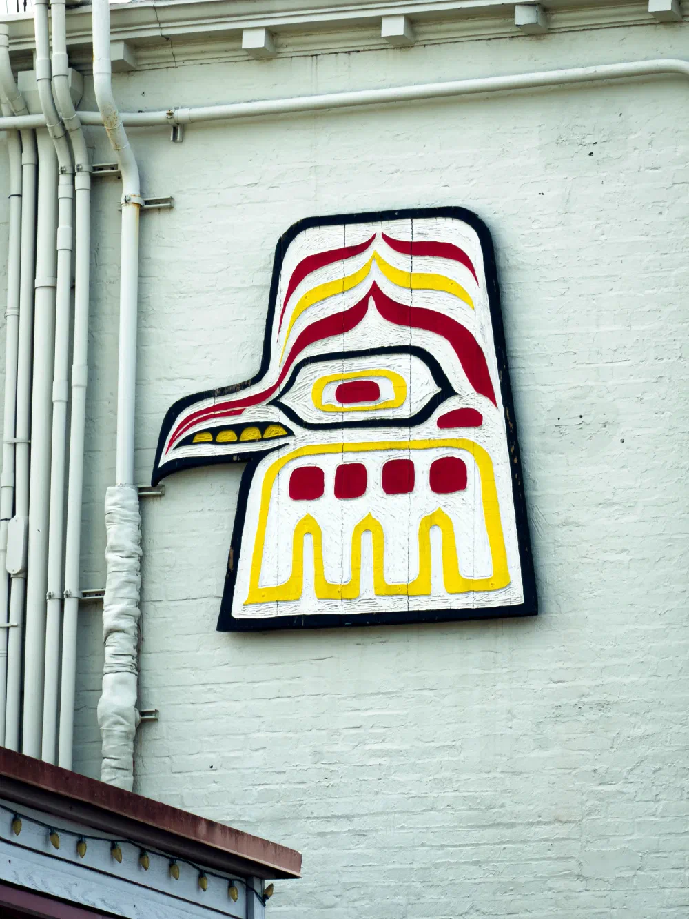 A stylized, colorful mural resembling a bird's head is painted on a white brick wall next to several vertical pipes.