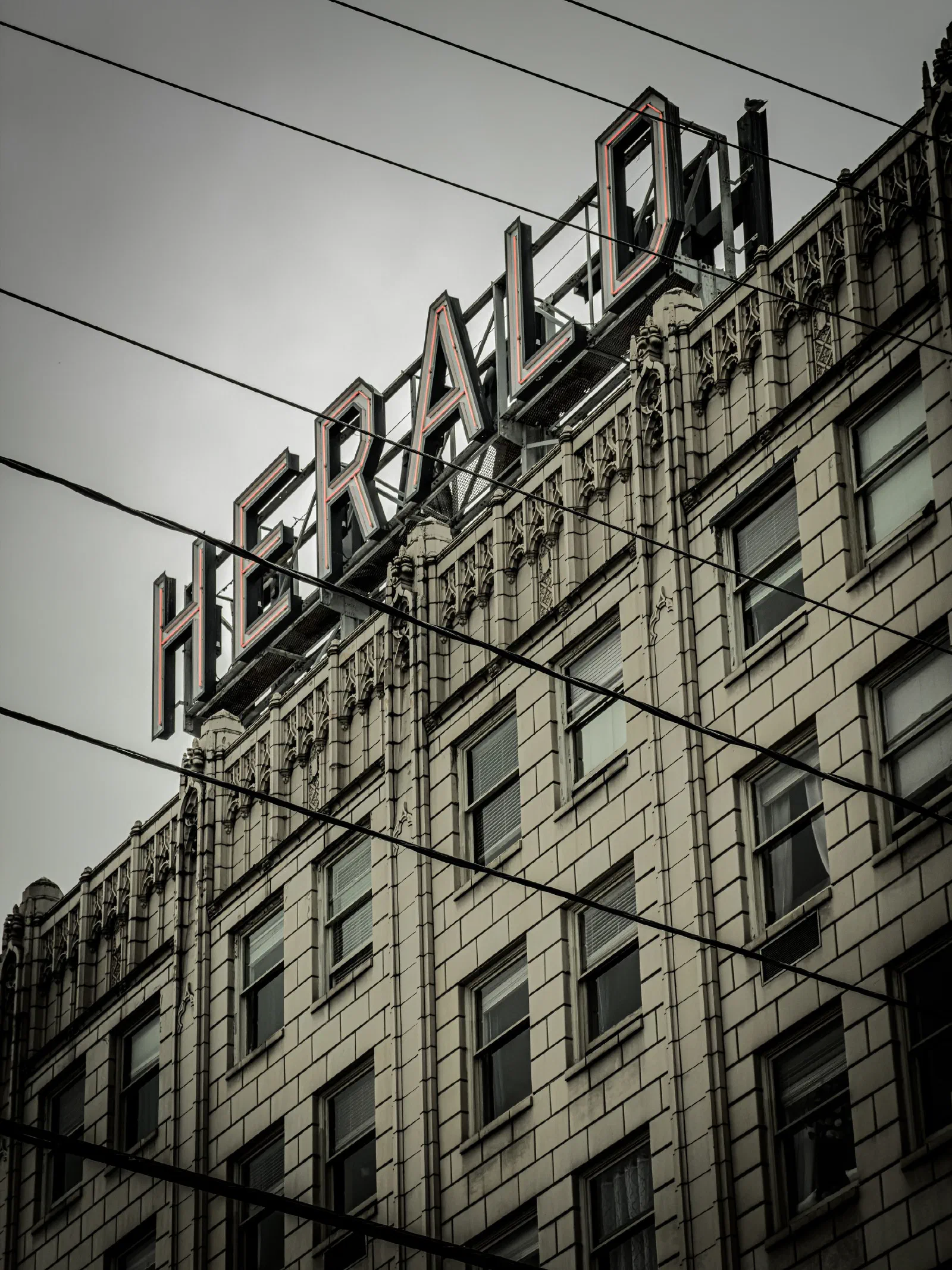 A tall, ornate building features a large HERALD sign on its roof with power lines crossing in front against a cloudy sky.