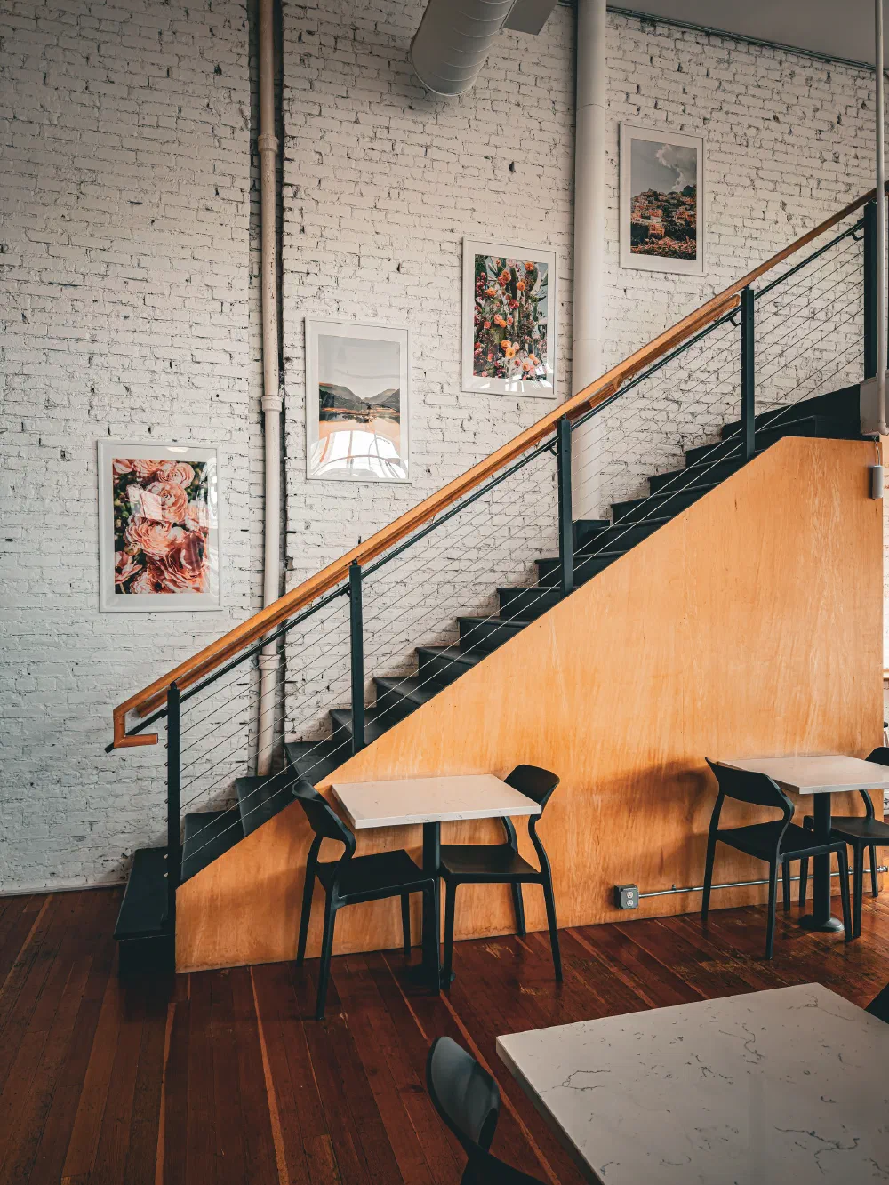 A cozy cafe interior features wooden floors, a staircase with a black railing and wooden paneling, and framed floral and nature artwork on a white brick wall.