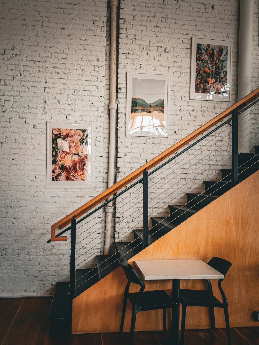 A small table with two black chairs is placed under a staircase decorated with three framed pictures on a white brick wall.