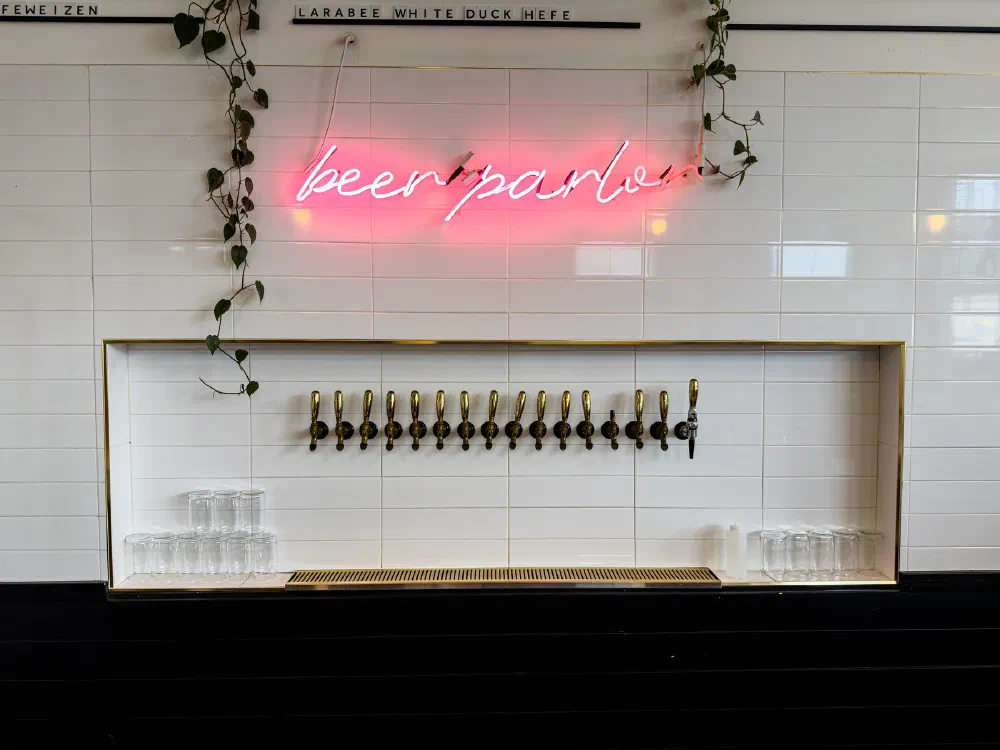 A row of beer taps is mounted on a white tiled wall with a pink neon sign reading beer parlor and vines hanging above.