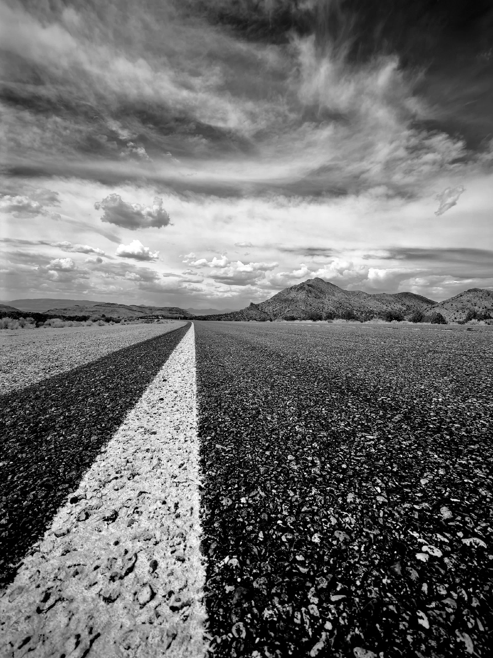 A long, empty road stretches toward distant mountains under a dramatic cloudy sky in black and white.